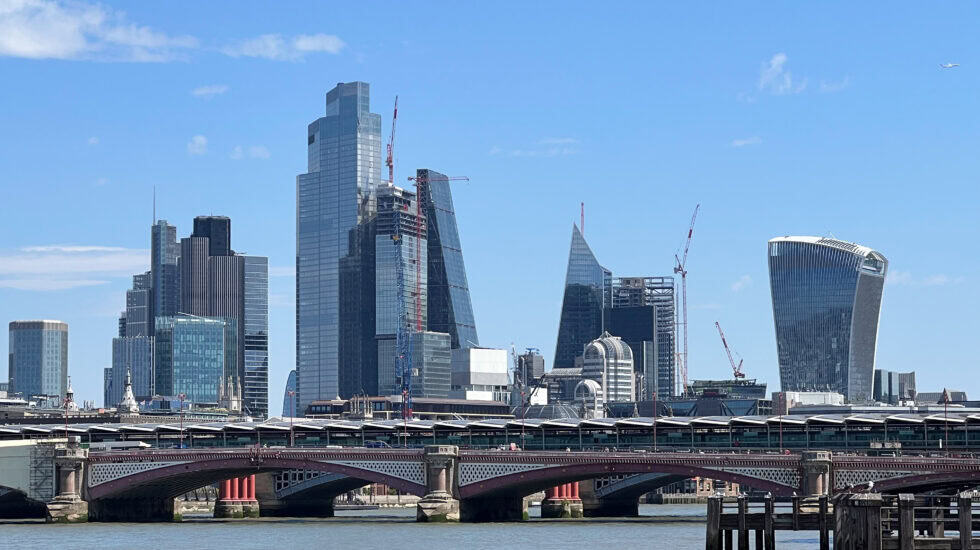 London skyline with modern skyscrapers including the Walkie Talkie and the Cheesegrater, viewed from across the River Thames under a clear blue sky.