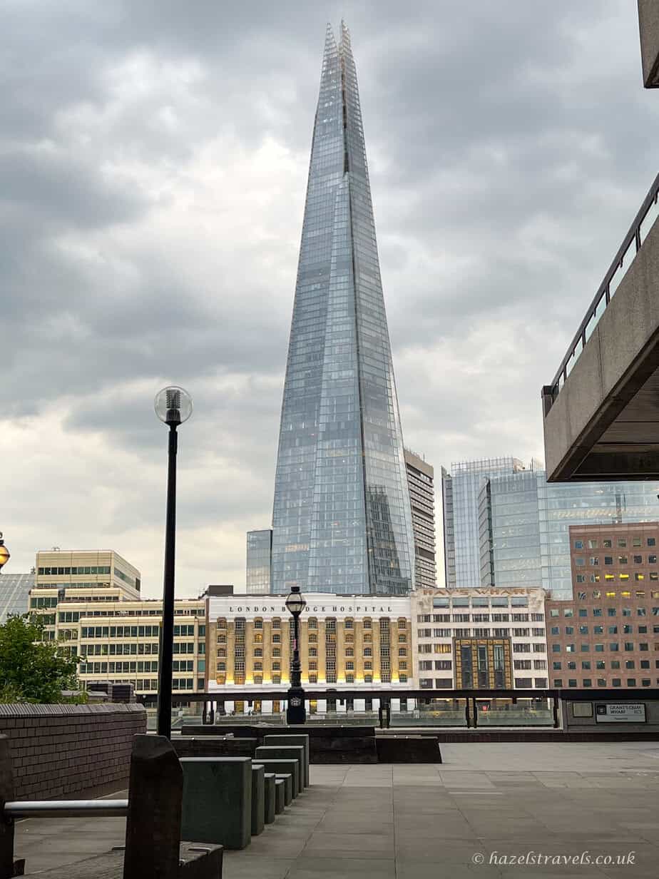 The Shard skyscraper in London rising into a pale blue sky with soft clouds, surrounded by modern buildings.