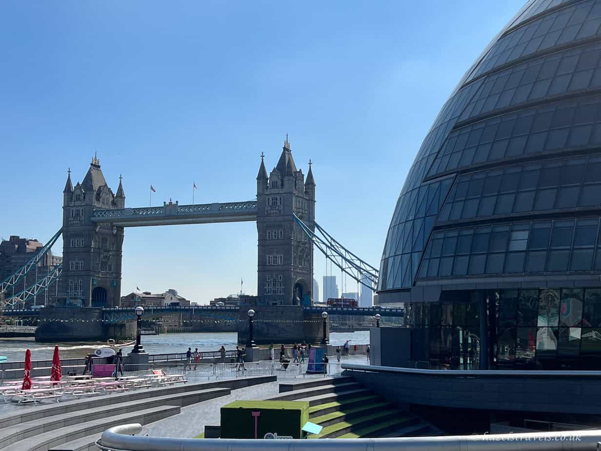 Tower Bridge and London’s curved glass City Hall building on a sunny day with bright blue sky.
