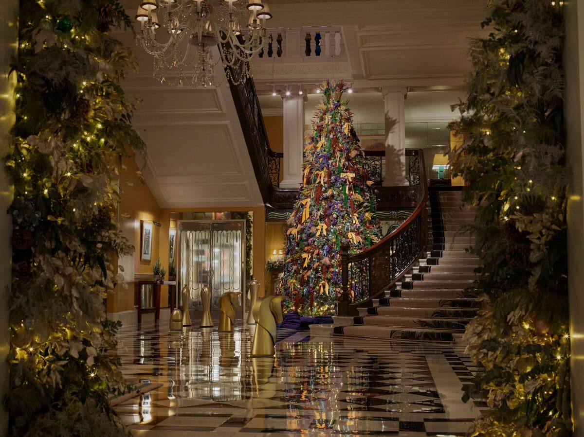 Lavishly decorated Christmas tree at Claridge’s in London, adorned with purple, gold and green ornaments, standing beside a sweeping marble staircase and reflected on a glossy black-and-white chequered floor in the hotel’s grand lobby.