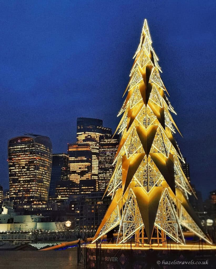 Illuminated modern Christmas tree in London glowing gold at night, set against a deep blue sky with the city skyline and skyscrapers in the background along the river