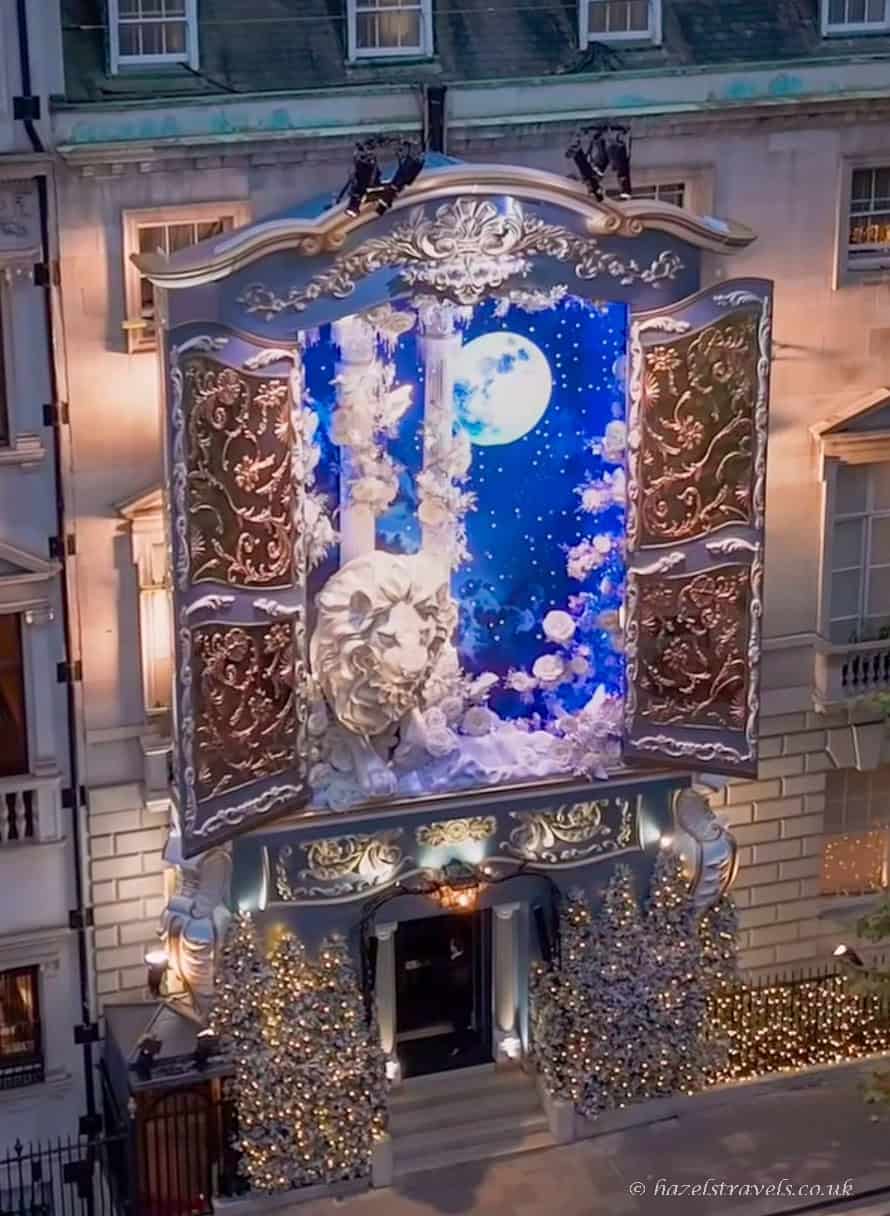 Lavish London townhouse Christmas display featuring a giant silver storybook façade with ornate gold detailing, a bright blue night-sky backdrop with a full moon, white roses, and a large sculpted lionâs head above sparkling silver Christmas trees.