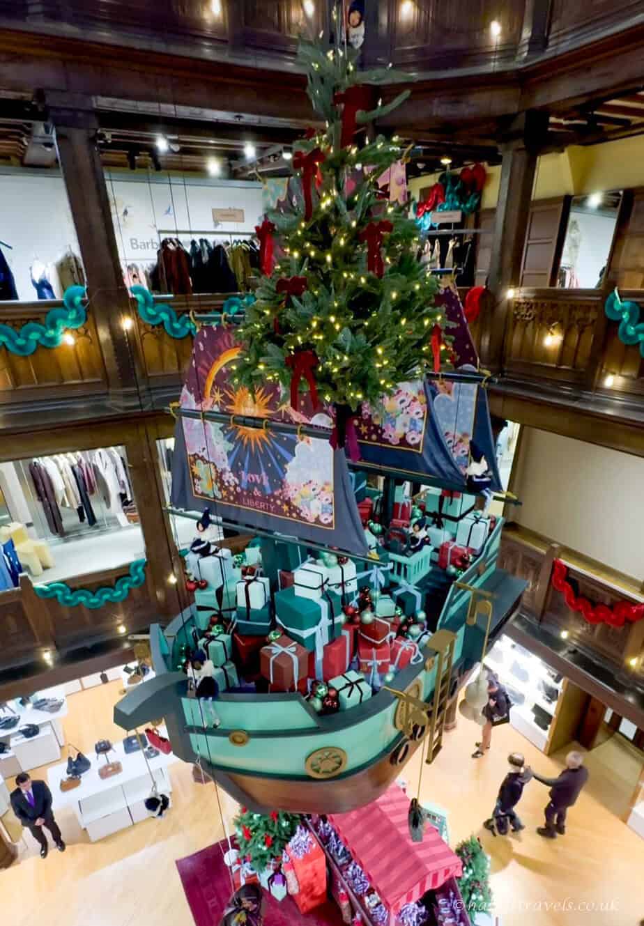 Liberty Londonâs Christmas display featuring a large turquoise pirate-style ship suspended in the atrium, filled with red, white, and teal gift boxes, with a decorated green Christmas tree rising from the centre and colourful patterned sails.