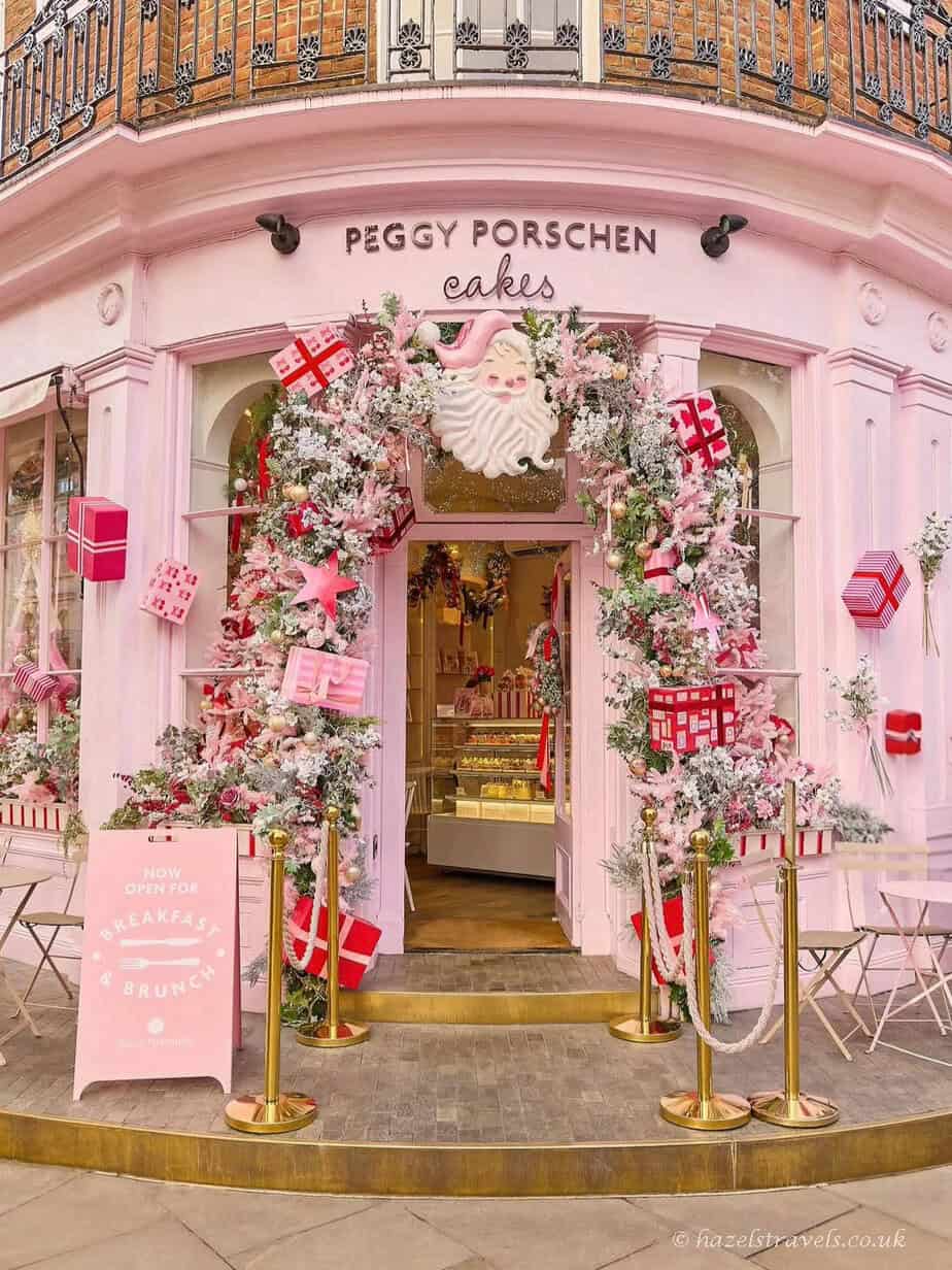 Peggy Porschen in Belgravia decorated for Christmas, featuring a pastel pink storefront framed with frosted white garlands, pink and red gift boxes, and a large white Santa face above the doorway.