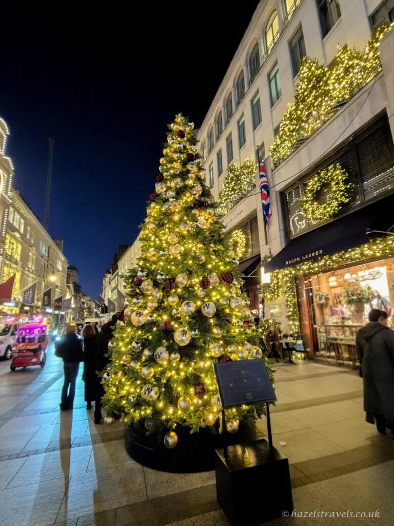 Large Christmas tree on New Bond Street decorated with warm golden lights, silver and gold baubles, and touches of red, surrounded by shopfronts covered in bright white and gold festive garlands at night.