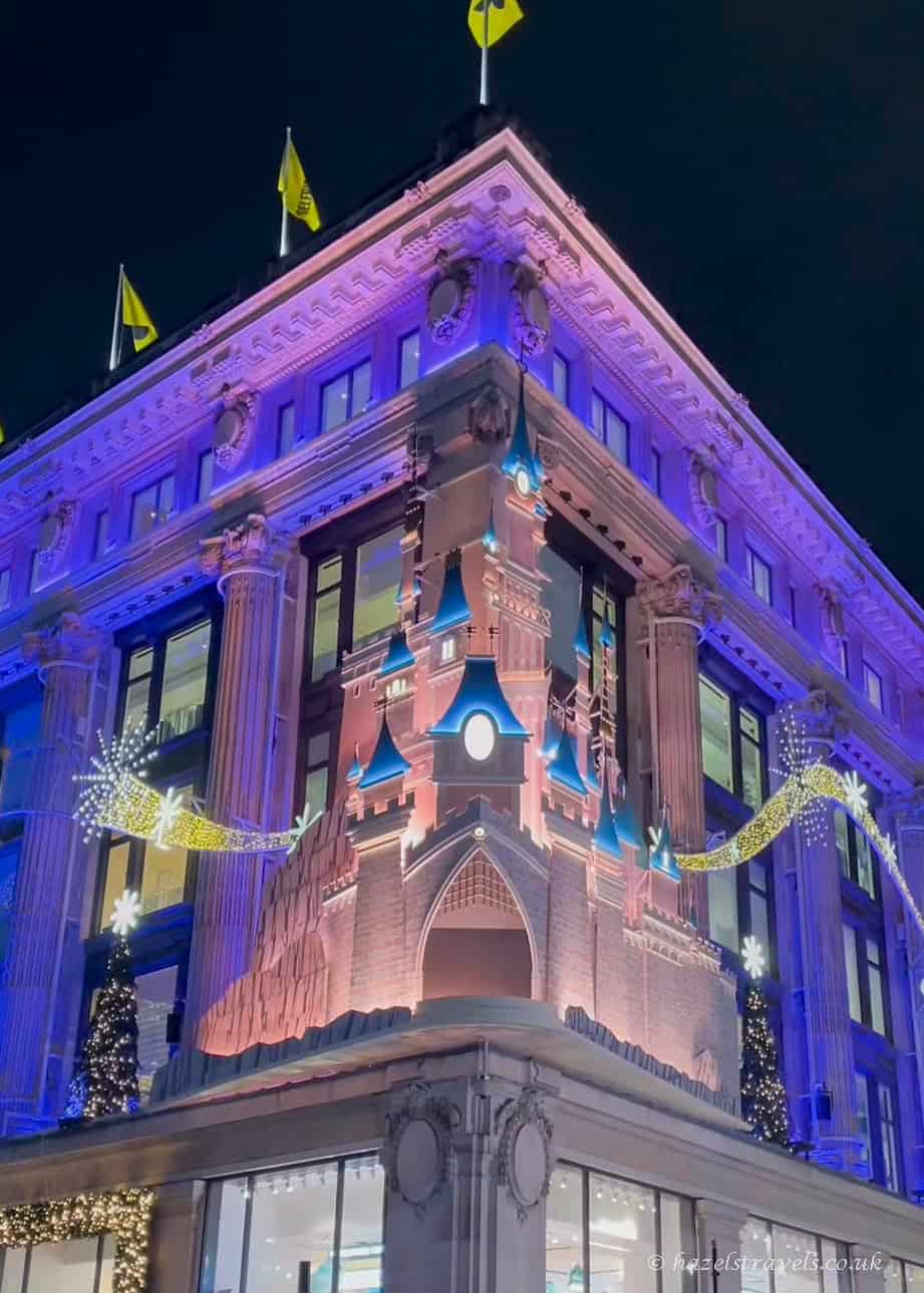 Disney-themed Christmas display on the corner of the Selfridges building, featuring a pink castle with bright blue turrets, golden shooting-star lights, and purple uplighting against the night sky.