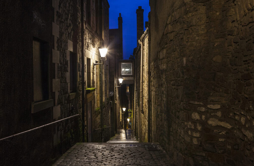 Narrow cobbled close in Edinburgh at night, with warm street lamps illuminating stone walls and steps leading downhill under a deep blue evening sky