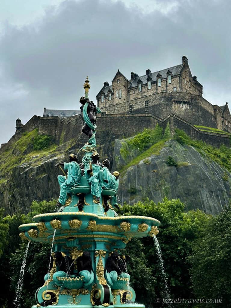 Ross Fountain in Princes Street Gardens with turquoise statues and flowing water, framed by Edinburgh Castle rising dramatically on the rocky hill behind under a cloudy sky