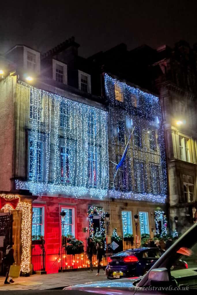 Georgian building on George Street in Edinburgh covered in cascading blue and white Christmas lights at night, with glowing windows, festive decorations, and cars passing in the foreground
