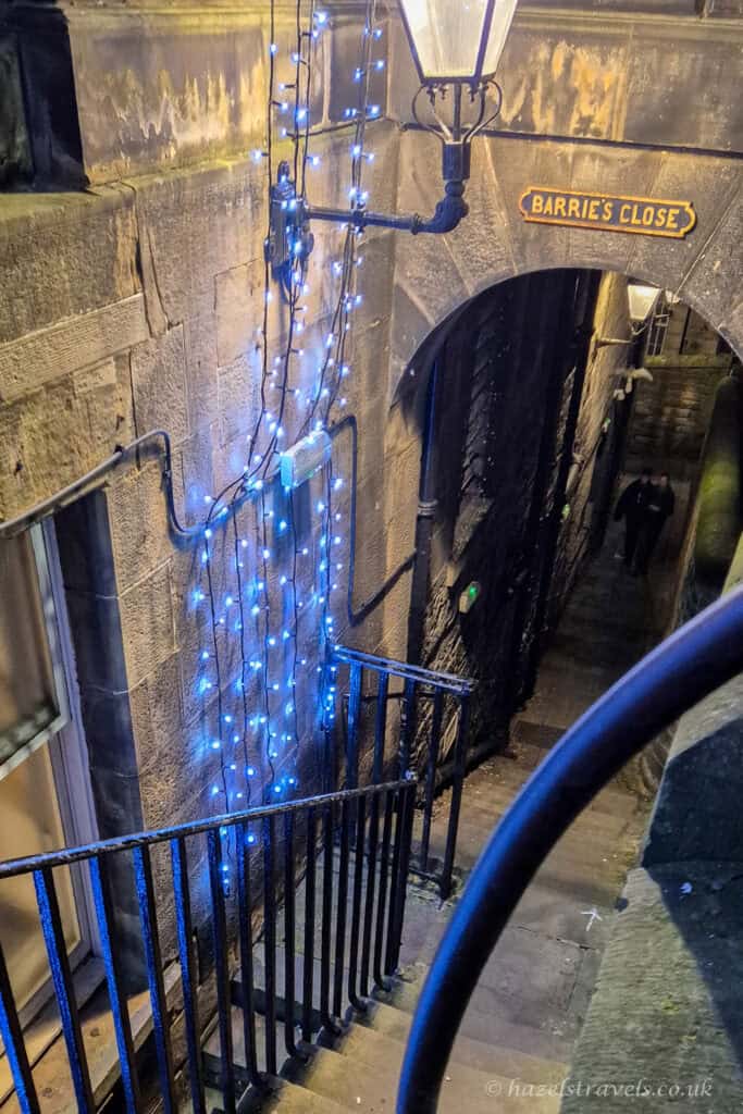 Stone steps leading into Barrie’s Close in Edinburgh, decorated with blue Christmas fairy lights, an old-fashioned lamppost, and a narrow arched passageway glowing at night