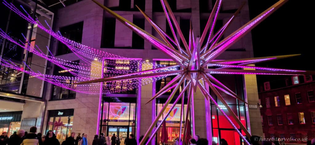 Large illuminated Christmas star installation glowing pink and purple at St James Quarter in Edinburgh, with festive light trails overhead and people gathered below at night
