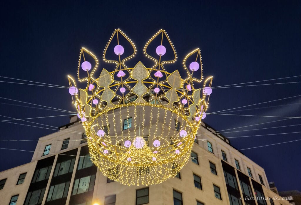 Giant golden crown Christmas light installation on Bond Street, decorated with glowing purple orbs and sparkling gold LEDs, suspended above the street against a deep navy evening sky.