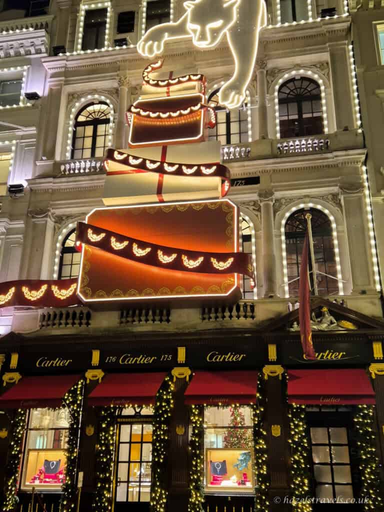 The Cartier store in London decorated for Christmas, featuring giant illuminated red-and-gold gift boxes stacked up the building with a glowing white panther above, festive garlands, and red awnings along the storefront.