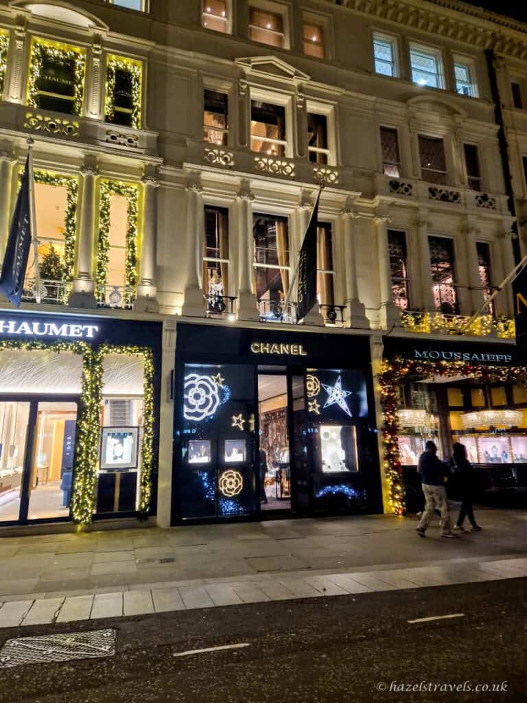 The Chanel boutique on Bond Street decorated for Christmas, with illuminated white and gold star and camellia lights on the storefront, and neighbouring luxury shops framed with glowing festive garlands.