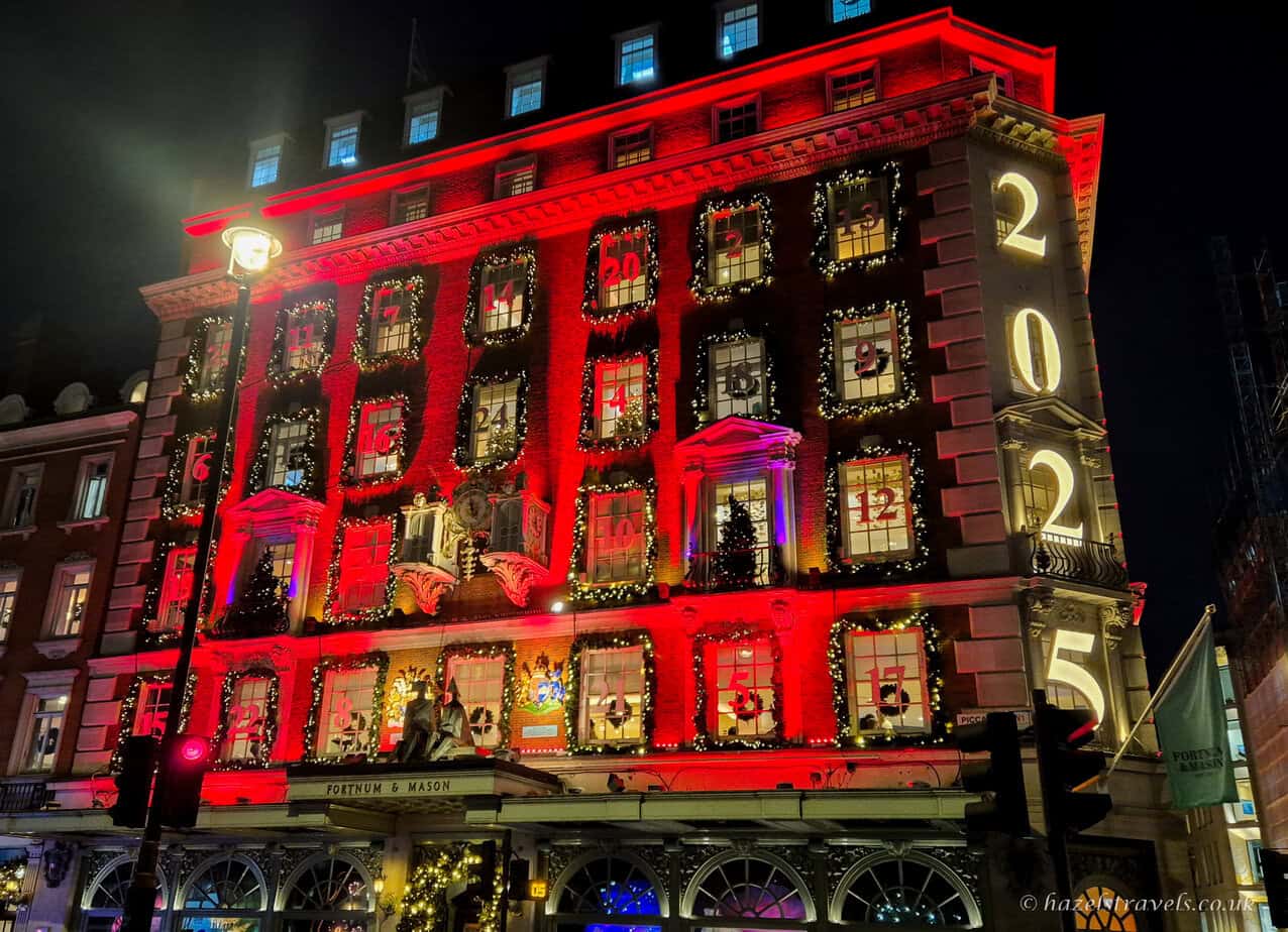 The Fortnum & Mason building in London illuminated in bright red for Christmas, with each window styled as an advent calendar and the year 2025 glowing vertically down the corner of the facade.