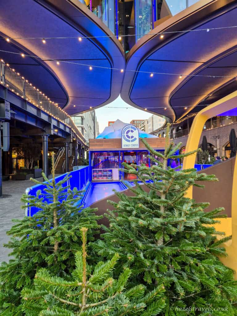 Outdoor winter bowling lane at King’s Cross, framed by small Christmas trees and modern illuminated architecture overhead, with vibrant blue lighting along the lanes.