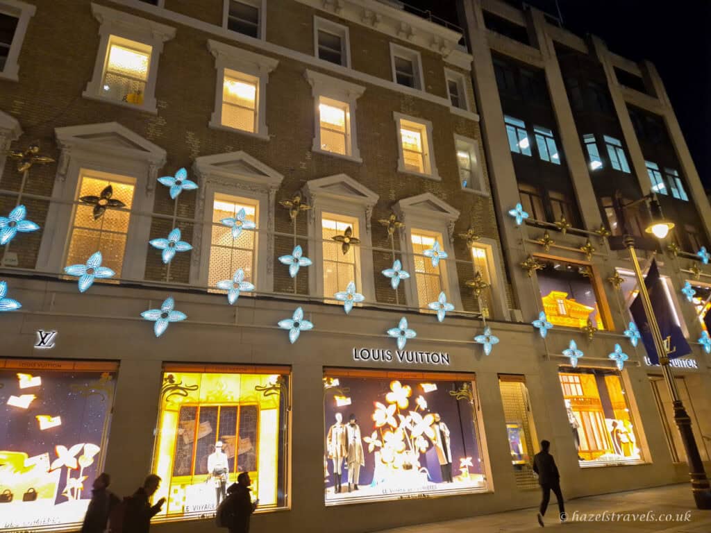 Louis Vuitton store on New Bond Street decorated with glowing blue and gold floral Christmas lights, with illuminated window displays and shoppers walking past at night.