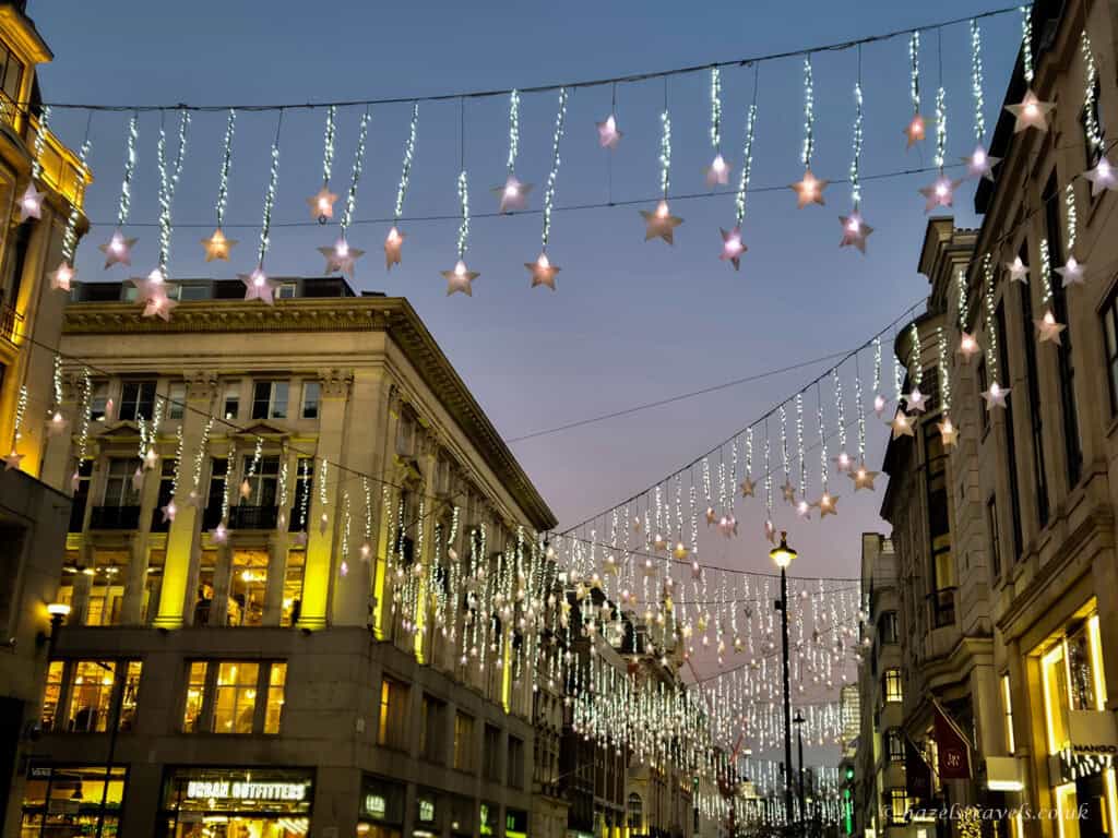 Star-shaped Christmas lights and white icicle garlands hanging above a London shopping street at dusk, with golden-lit buildings lining the scene.