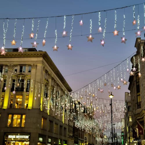 Star-shaped Christmas lights and white icicle garlands hanging above a London shopping street at dusk, with golden-lit buildings lining the scene.