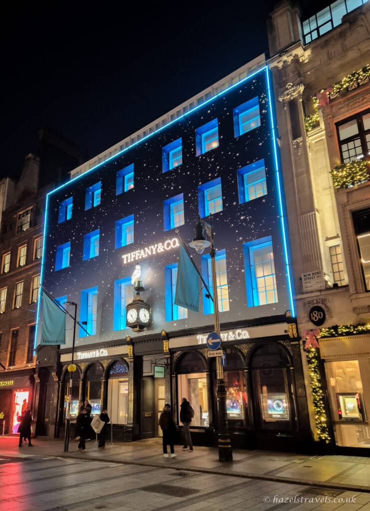 Tiffany & Co. store on Bond Street illuminated with bright blue Christmas lights, glowing window displays and a starry night-themed façade, with shoppers walking past under the dark evening sky.