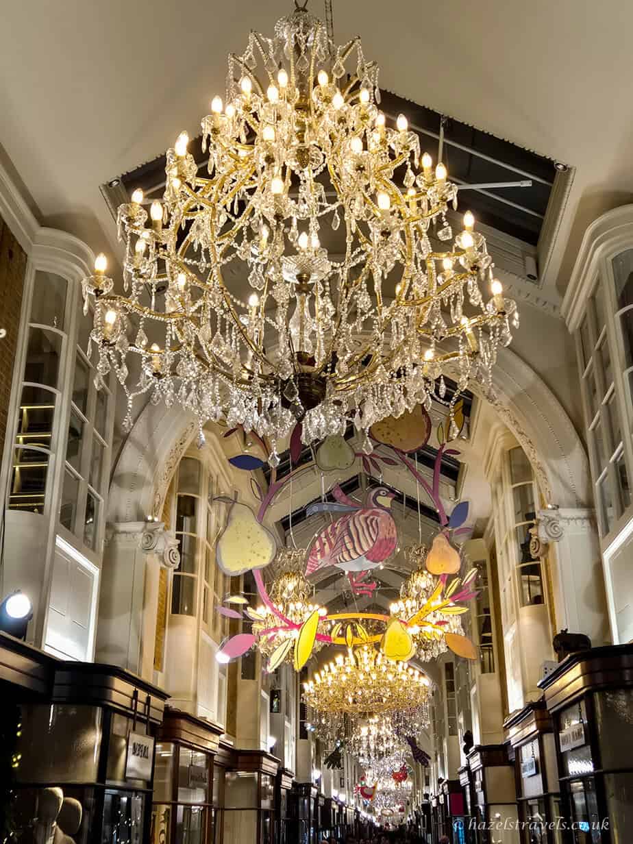 Sparkling golden chandeliers hanging above Burlington Arcade, with pastel Christmas decorations in pink, yellow, and purple suspended along the elegant cream-coloured corridor.