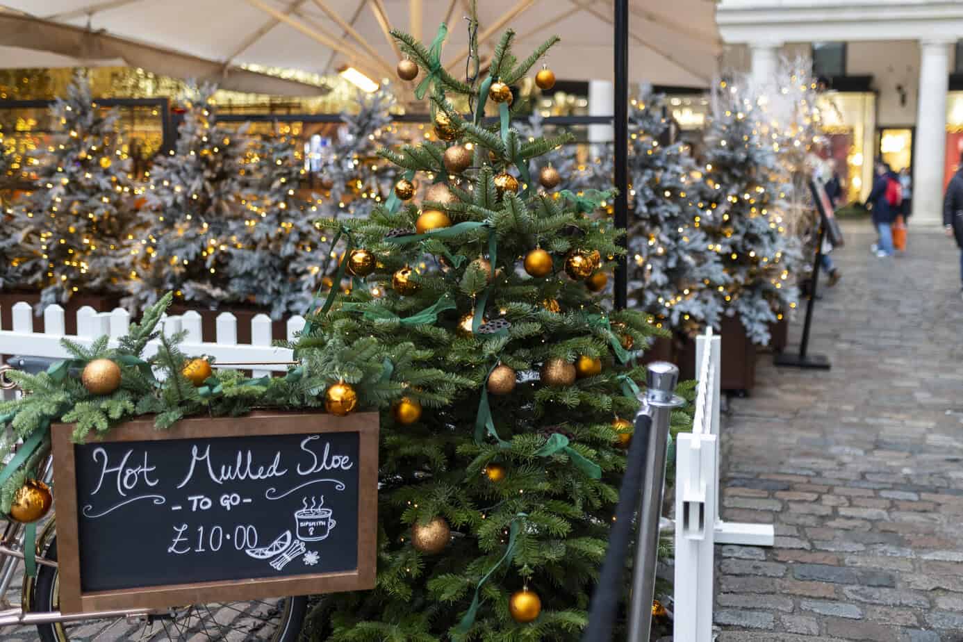 Christmas tree with gold baubles outside a mulled wine stall in Covent Garden, London, surrounded by snowy artificial trees and festive lights.