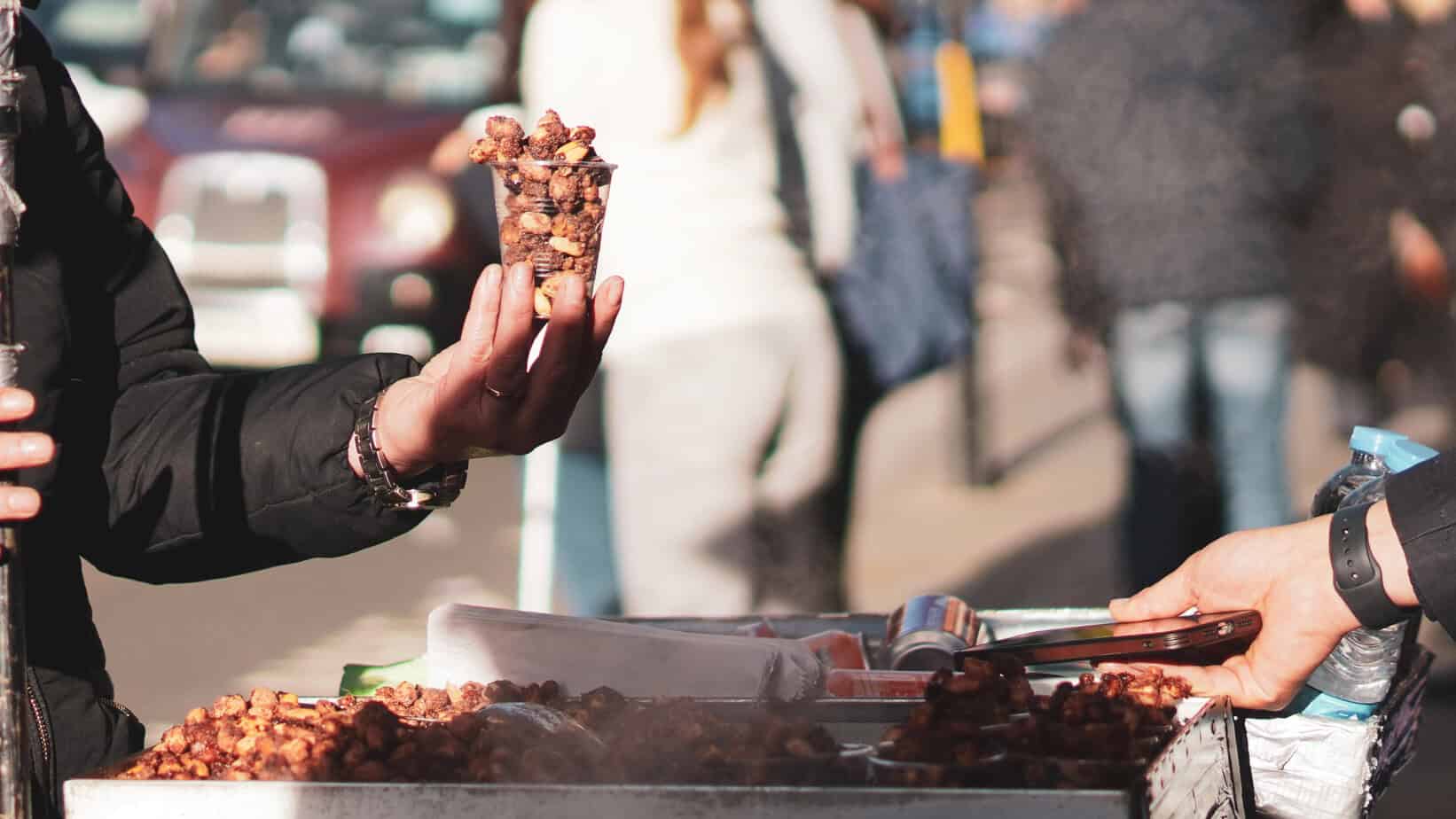 Street vendor holding a cone of roasted chestnuts at Greenwich Christmas Market in London, with blurred festive crowds in the background.