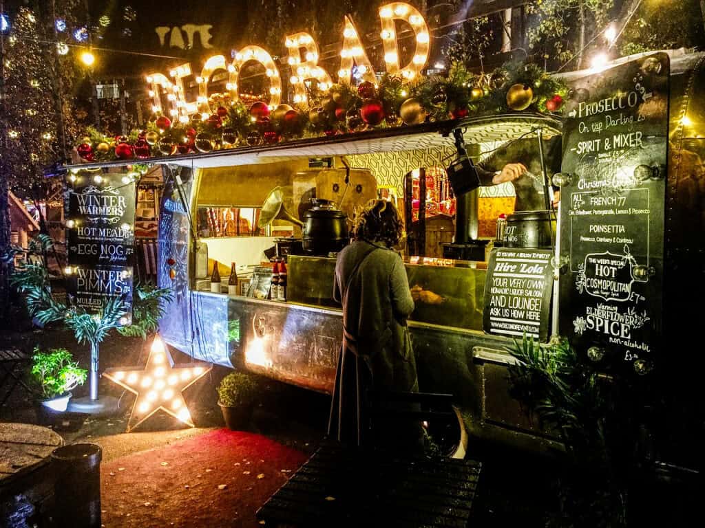 Festive South Bank Christmas market stall lit with warm golden lights, featuring a silver food truck decorated with red and gold baubles, green garlands, and a glowing star, as a customer in a grey coat orders drinks at night.