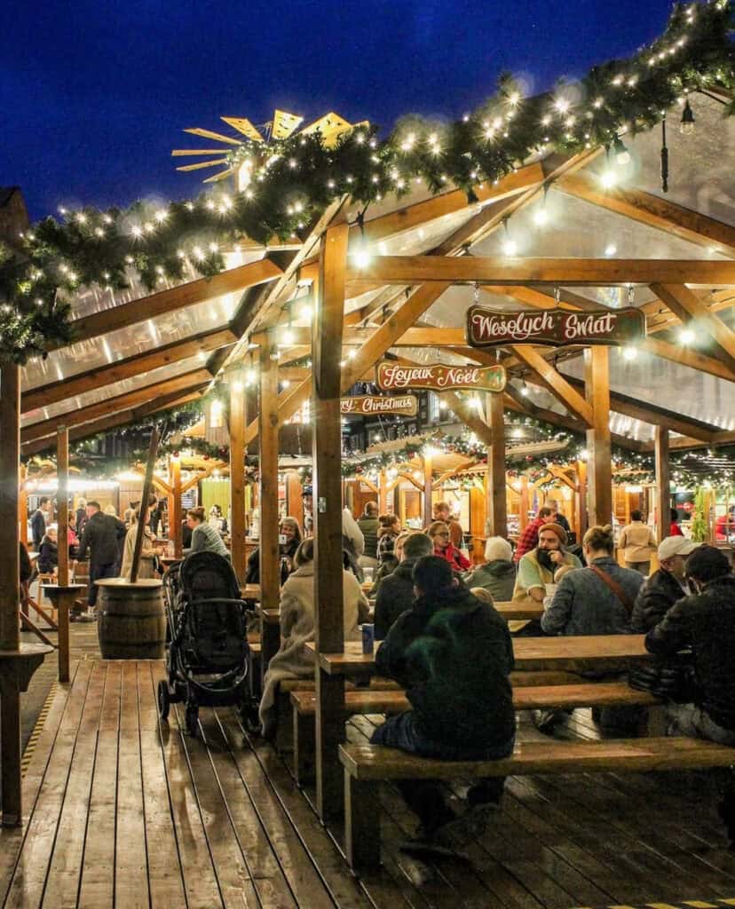 Kingston Christmas market with wooden chalet stalls and long communal tables, people sitting and chatting under warm fairy lights and garlands, festive signs above the stalls and a deep blue winter sky overhead