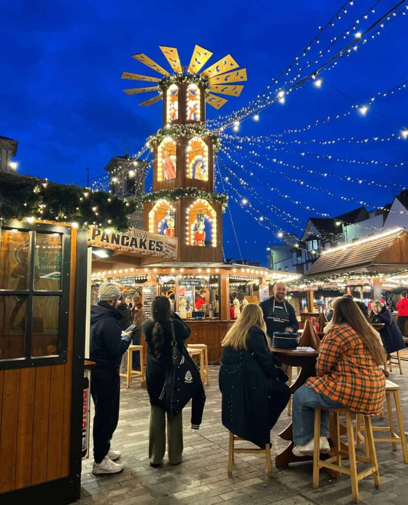 Kingston Christmas market at night with wooden festive stalls, people sitting and standing around high tables, warm string lights overhead and a tall illuminated Christmas pyramid against a deep blue winter sky