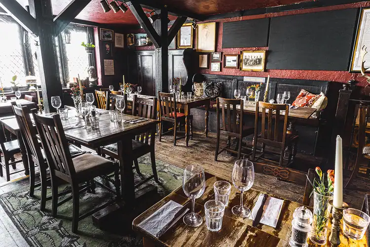 The cosy interior of The Mayflower pub in London, featuring rustic wooden tables set with wine glasses, vintage wooden chairs, dark timber beams, and warm, old-world décor with framed pictures and cushions along the bench seating.