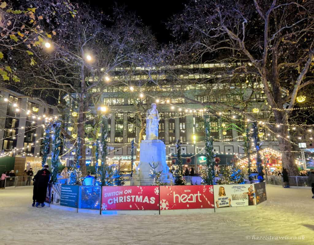 Leicester Square’s illuminated Christmas ice rink at night, with twinkling fairy lights strung through tall trees, colourful festive banners around the central statue, and people skating beneath the bright city backdrop.
