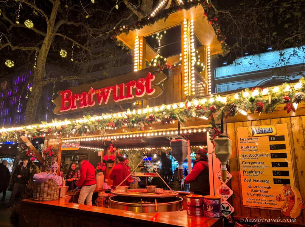 Festive bratwurst stall at Leicester Square Christmas Market, decorated with glowing fairy lights, red garlands and pine branches, with vendors grilling sausages behind the counter under a warm golden evening glow.