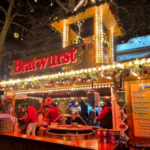 Festive bratwurst stall at Leicester Square Christmas Market, decorated with glowing fairy lights, red garlands and pine branches, with vendors grilling sausages behind the counter under a warm golden evening glow.