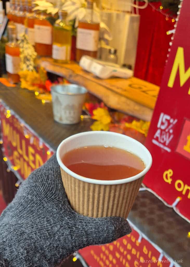 Hot spiced mulled gin in a brown paper cup at the Edinburgh Christmas Market, held in a gloved hand with festive stall decorations in the background