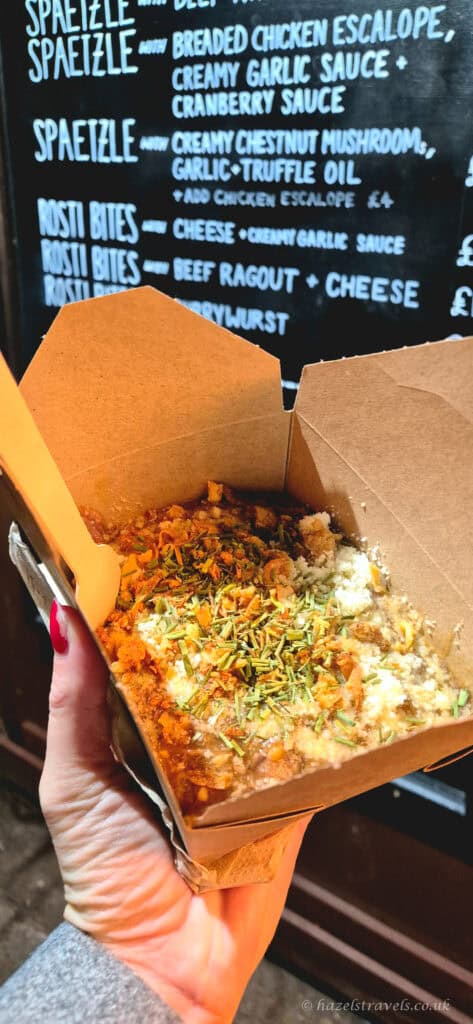Spaetzle with beef ragout and melted cheese from the Edinburgh Christmas Market, served in a cardboard tray with herbs on top and menu board in the background