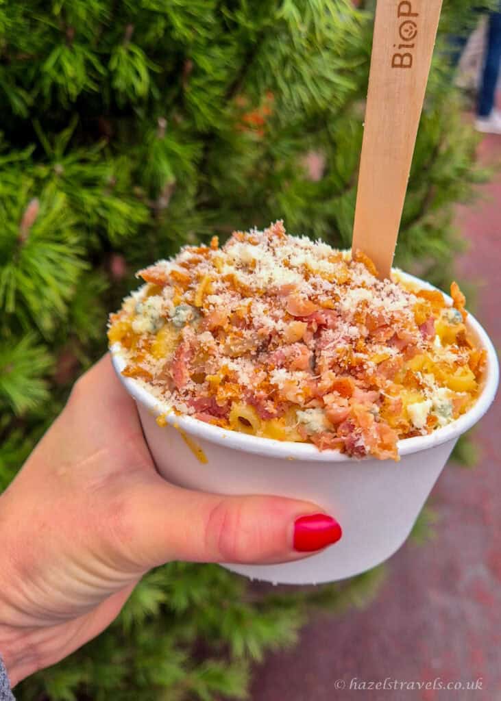 Mac and cheese topped with bacon and blue cheese at the Edinburgh Christmas Market, served in a white paper bowl with a wooden spoon