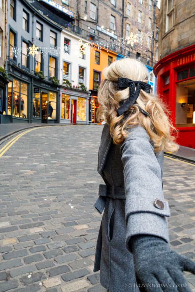 Woman walking along Victoria Street in Edinburgh during the festive season, with colourful shopfronts, Christmas lights and cobbled street