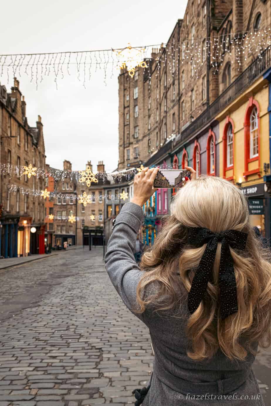 Woman taking a photo on Victoria Street in Edinburgh during the festive season, with Christmas lights and colourful buildings lining the cobbled street