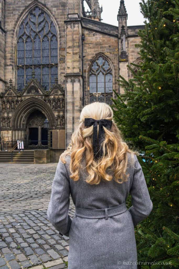 Woman standing near a Christmas tree outside St Giles’ Cathedral in Edinburgh, with historic stone architecture and cobbled courtyar