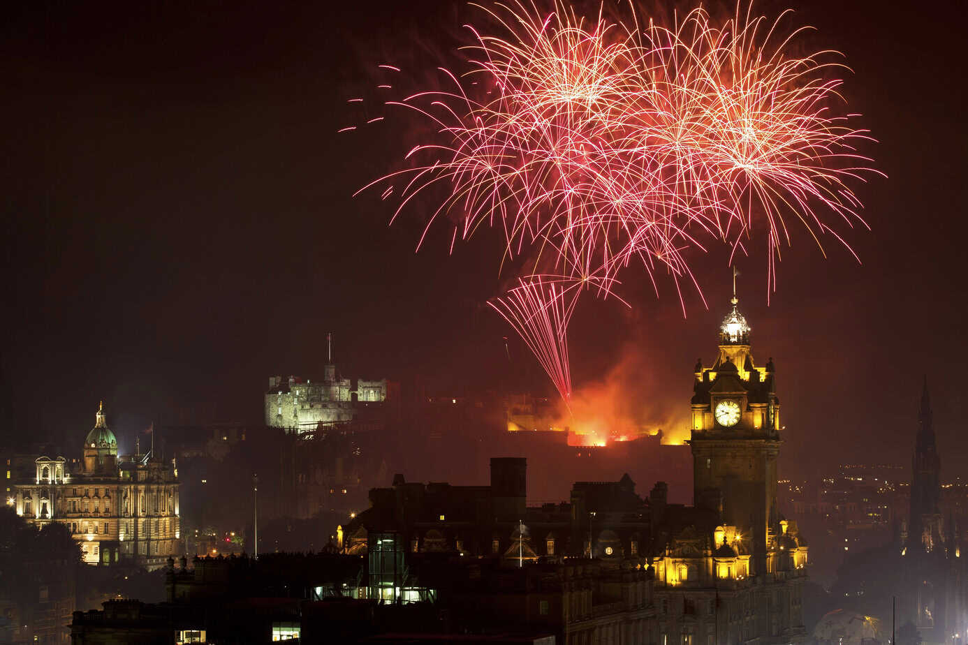 Red fireworks lighting up the sky above Edinburgh Castle during Hogmanay celebrations, with historic buildings and the Balmoral Clock Tower in the foreground