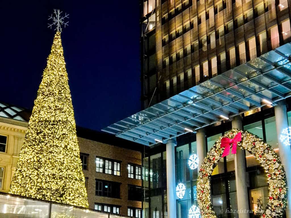 Tall Christmas tree covered in warm golden lights with a glowing star topper, standing beside a festive illuminated arch with a red bow outside a modern glass building at night, under a deep navy blue winter sky in London
