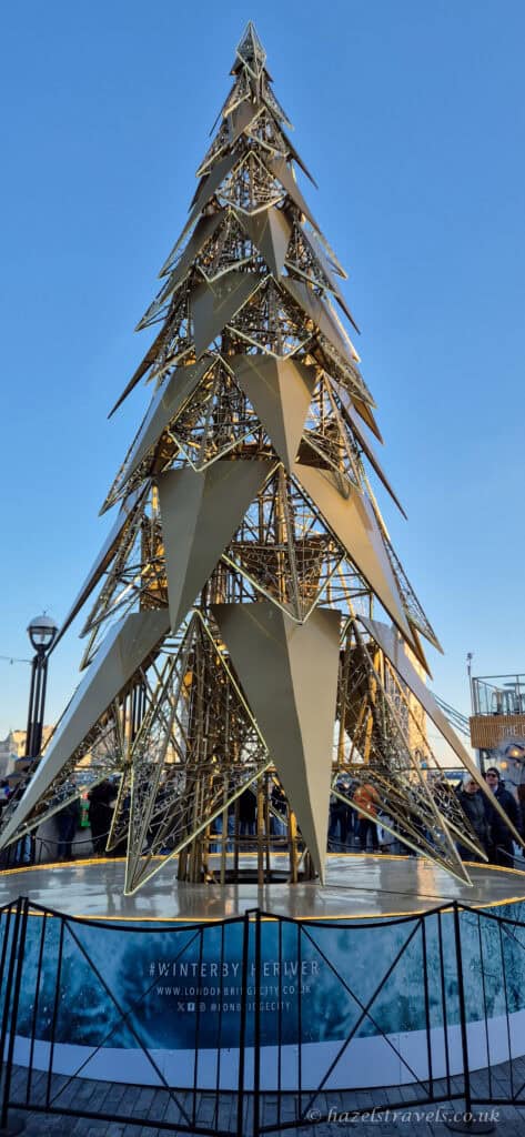 Modern sculptural Christmas tree made from gold metal panels at Winter by the River on London’s South Bank, set against a clear bright blue winter sky with people gathered around the base