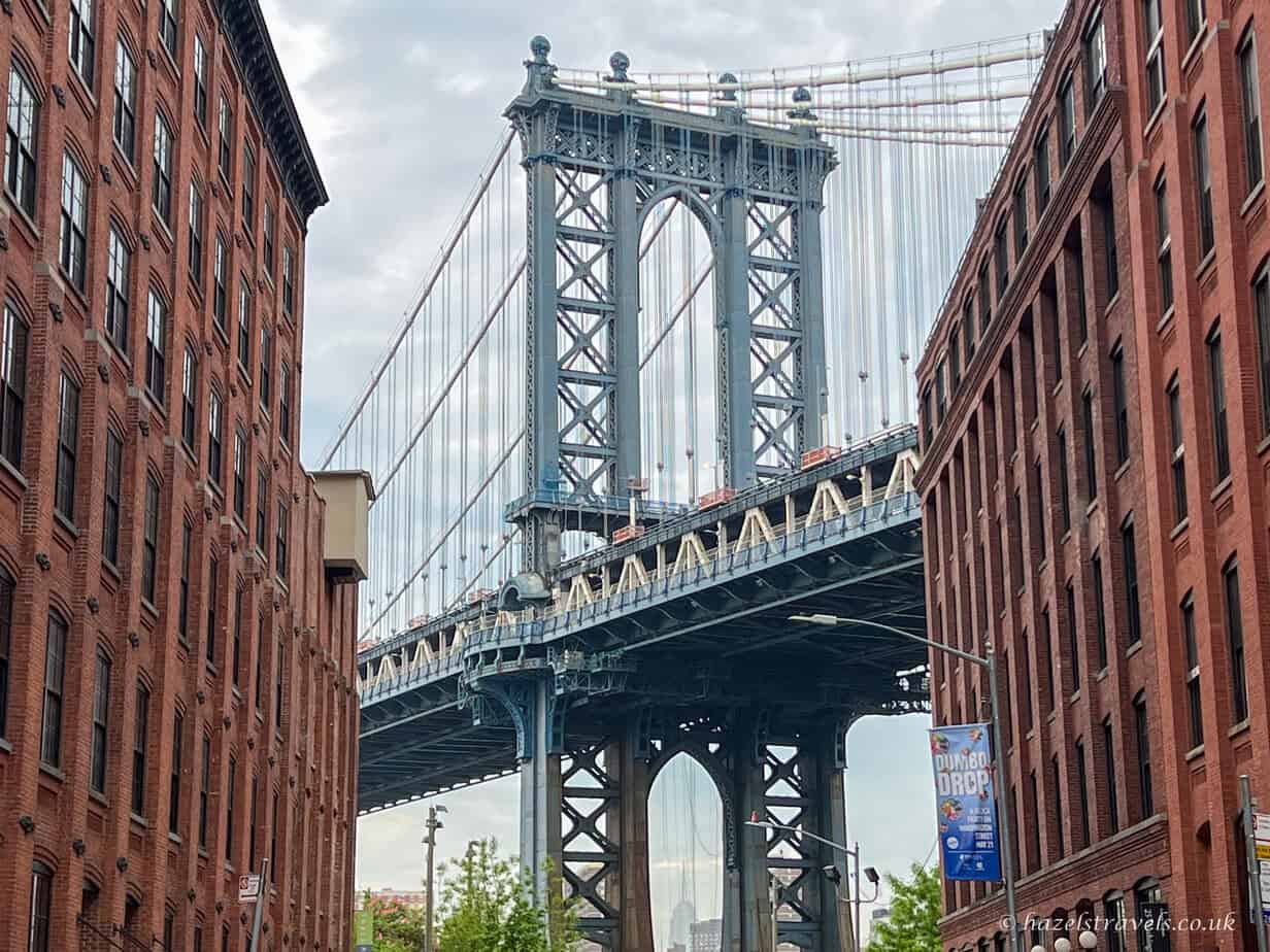 Manhattan Bridge framed between red brick buildings in DUMBO, Brooklyn, with steel towers and suspension cables rising above the cobbled street