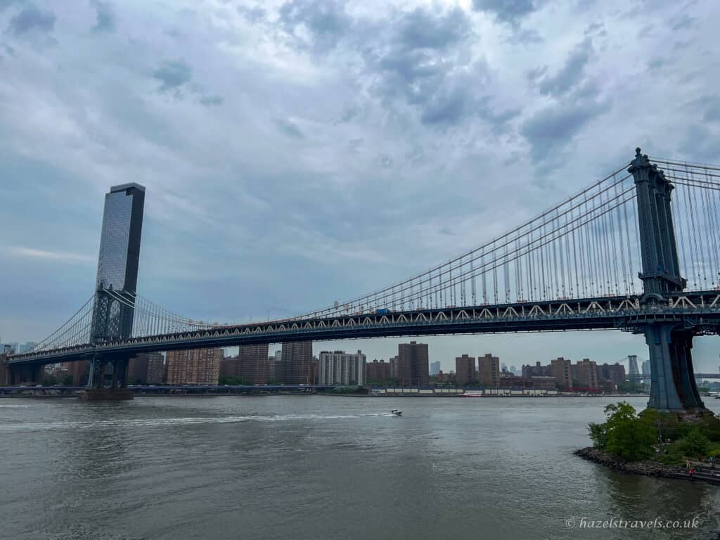 Manhattan Bridge spanning the East River in New York City, with steel suspension cables, river traffic below, and a cloudy sky above