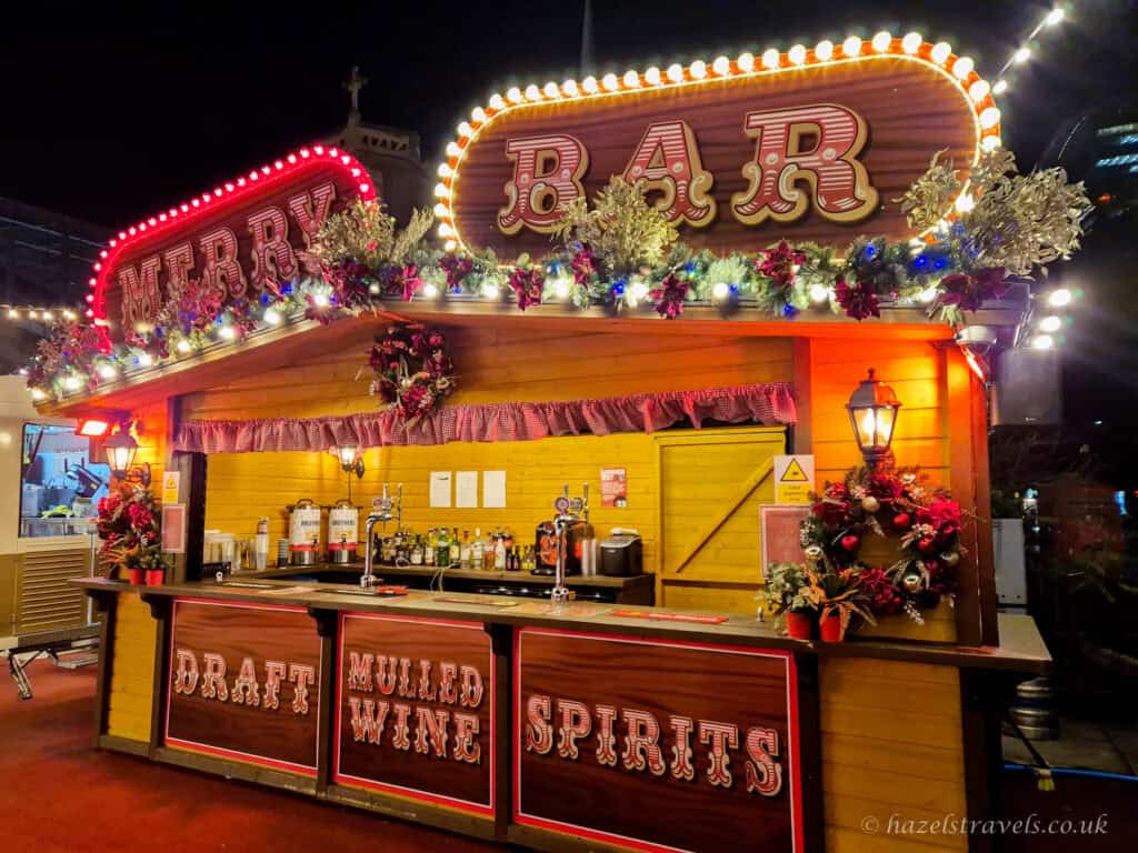 Brightly lit Christmas market bar stall with red and white BAR and MERRY signs outlined in warm bulbs, yellow wooden walls decorated with green garlands and red baubles, and counters labelled Draft, Mulled Wine and Spirits glowing against a dark night sky in London