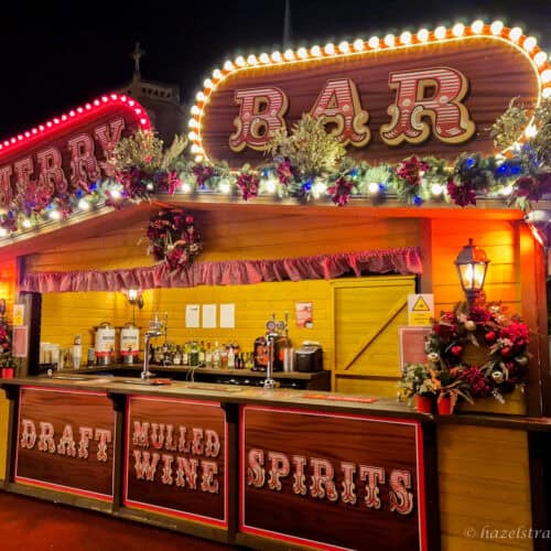 Brightly lit Christmas market bar stall with red and white BAR and MERRY signs outlined in warm bulbs, yellow wooden walls decorated with green garlands and red baubles, and counters labelled Draft, Mulled Wine and Spirits glowing against a dark night sky in London