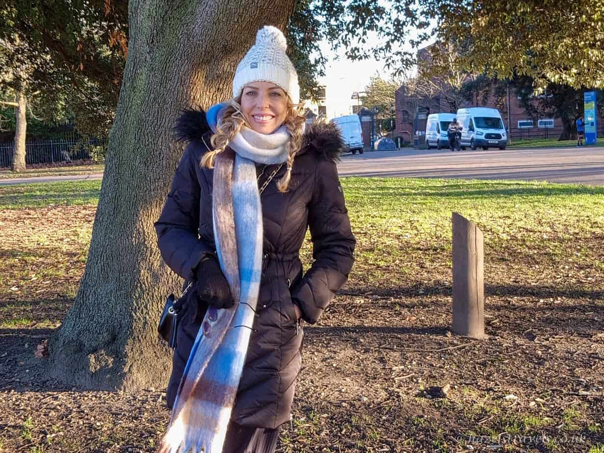 Smiling woman in a winter coat, knitted hat and long striped scarf standing by a tree in Victoria Park, Hackney, on a bright winter afternoon