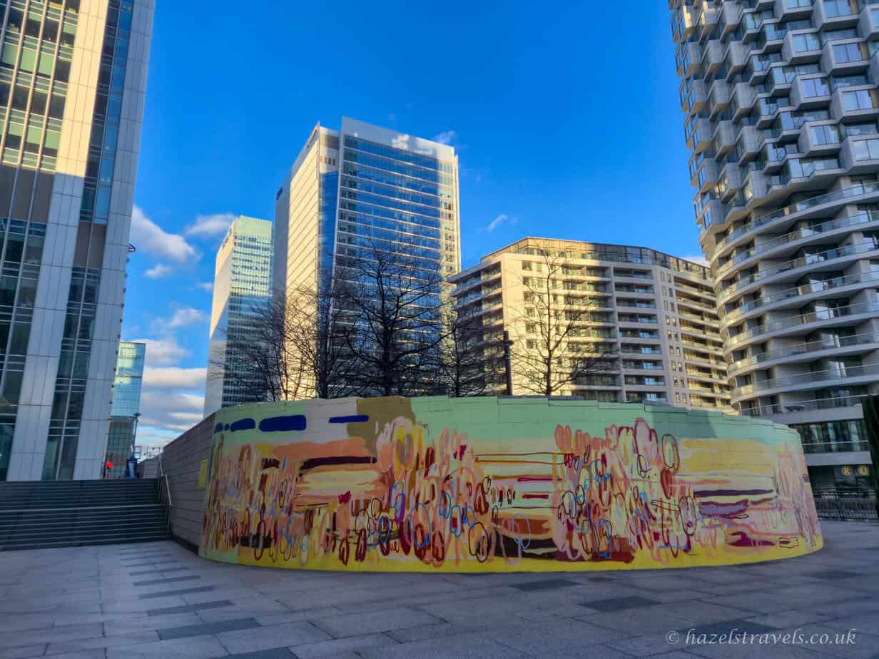 Colourful abstract street art mural in Canary Wharf, London, with modern glass skyscrapers and curved apartment buildings rising behind it under a bright blue sky