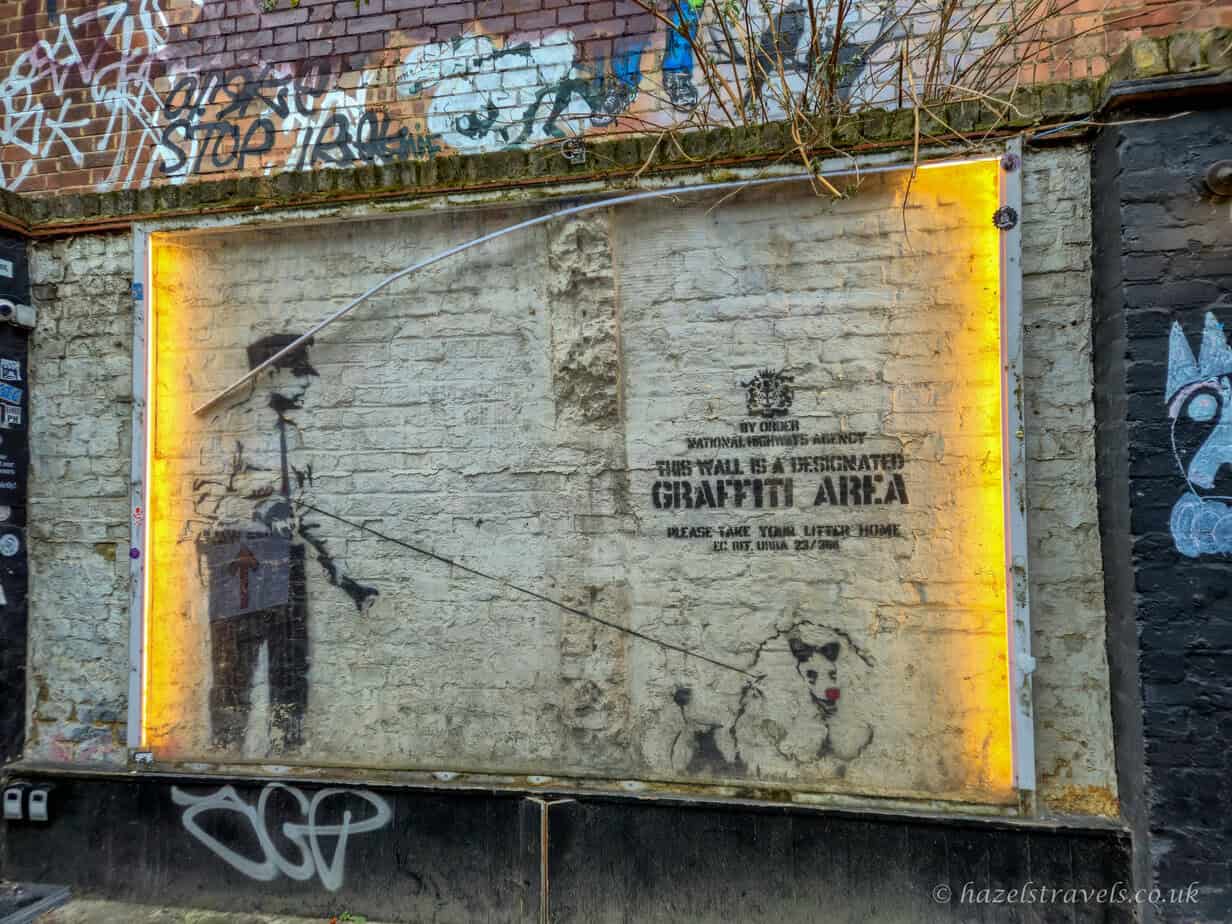 Banksy street art piece in Shoreditch, London, showing a stencilled figure and faded graffiti on a weathered concrete wall framed by a glowing yellow border